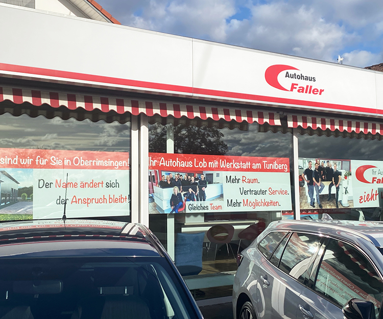 Breites Schaufenster mit mehreren Schildern für die Umzugswerbung des Lob Autohaus. Davor stehen Autos.